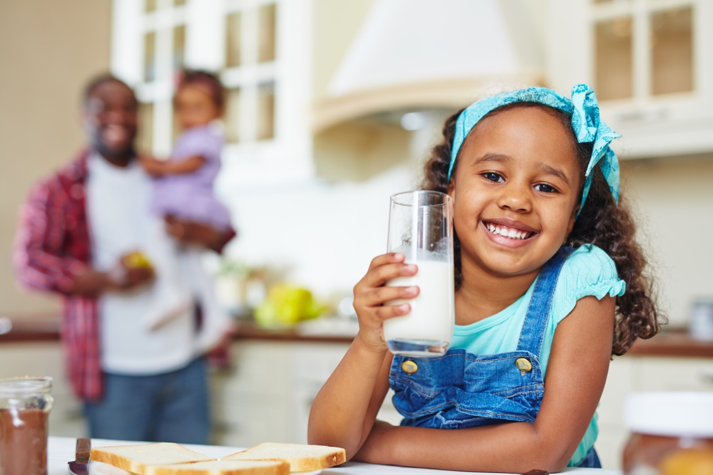 Happy African-American girl holding a glass of milk rich in vitamin A, with her father and sister in the background.
